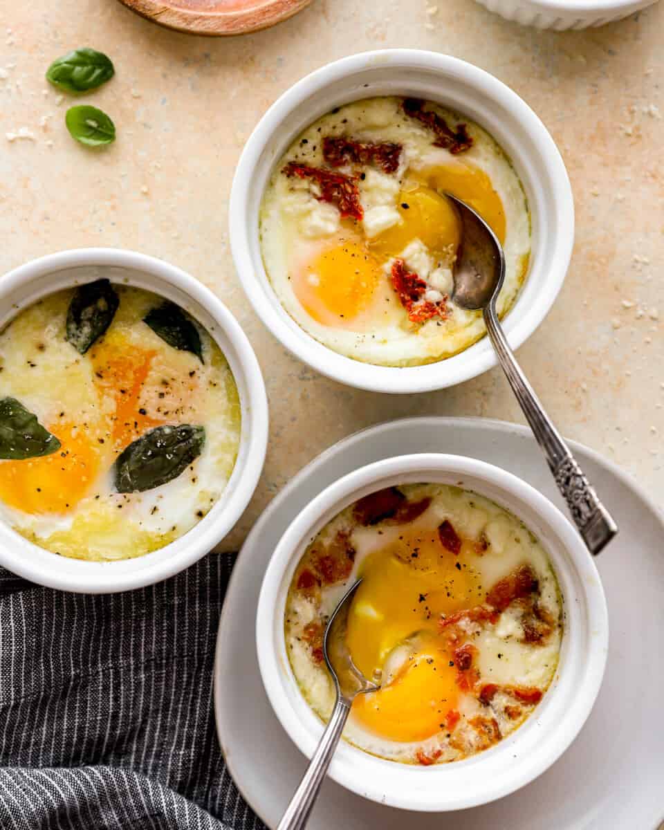 Three bowls of eggs with bacon and basil on a table.