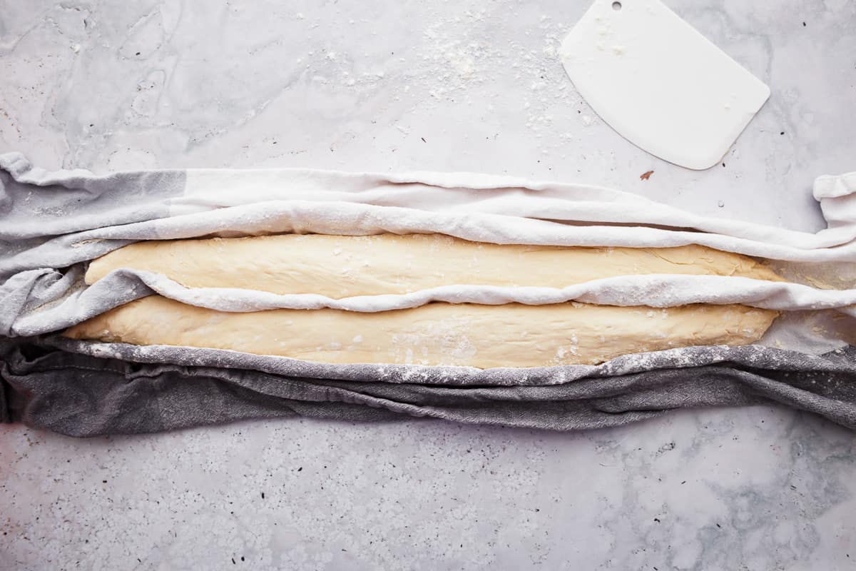 Long pieces of bread dough shaped into baguettes, sitting between folds of a dish towel while rising.